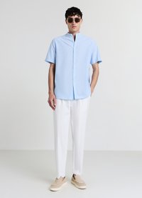 Chemise bleu clair à manches courtes avec boutons et col officier, associée à un pantalon blanc et des chaussures beige à enfiler. Tissu lisse et texturé.