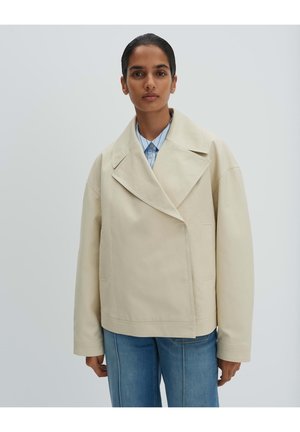 Person mit dunklen Haaren, die eine cremefarbene übergroße Jacke über einem blauen Hemd mit Kragen und blauer Jeans trägt, steht vor einem hellgrauen Hintergrund.