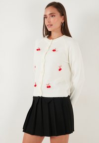 Witte gebreide cardigan met rode kersversieringen, ronde halslijn en knoopsluiting. Gecombineerd met een zwarte plooienrok.