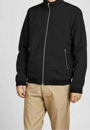 Man draagt een zwart jack met ritszakken en beige broek, staand tegen een effen lichte achtergrond.