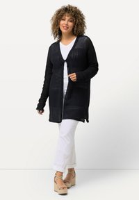 Svart stickad cardigan med öppen front, texturerat mönster och knytning fram. Bärs över en vit topp och kombineras med vita byxor och plattformssandaler.