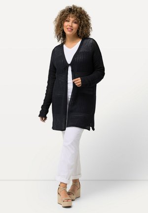 Cárdigan negro de punto con un frente abierto, patrón texturizado y lazo delantero. Lleva puesto sobre una blusa blanca y combinado con pantalones blancos y sandalias de plataforma.