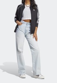 Kvinna klädd i svart Adidas-jacka, vit kortärmad perforerad topp, ljusblå vida jeans, vita sneakers och guldhalskedja.