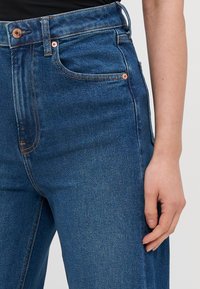 Mörkblå högmidjade jeans i denim. Har kopparfärgad hårdvara, framfickor och synlig sömnad längs sömmarna.
