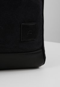 Sac à dos rectangulaire en tissu noir avec une surface texturée, doté d'une base en cuir noir lisse et d'une petite étiquette en relief.