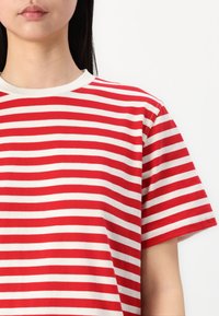 Γυναίκα που φοράει μπλουζάκι με κόκκινες και λευκές οριζόντιες ρίγες, κοντά μανίκια και στρογγυλή λαιμόκοψη, σε απλό λευκό φόντο.