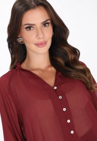 Pitkäaikaisilla aaltoilevilla ruskeilla hiuksilla varustettu nainen käyttää suuria geometrisia korvakoruja, herkkää kultakaulakorua ja burgundinpunaisen napitettua puseroa, jossa on läpikuultavaa kangasta.