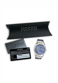 Montre en argent avec un cadran rond irisé bleu et des aiguilles argentées, dotée d'un bracelet à motif multicolore, emballée avec une carte de garantie noire.