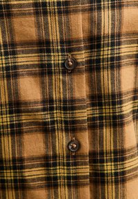 Camisa de cuadros marrón y amarilla hecha de franela, con líneas negras, textura de tela y botones marrones en la parte delantera.