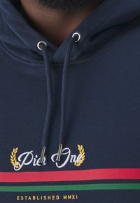 Sudadera azul marino de tela suave, con un logo bordado, rayas horizontales rojas y verdes, y detalles metálicos en los cordones.