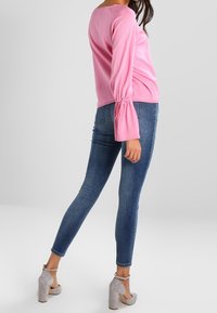 Top rosa a maniche lunghe con polsini arricciati, abbinato a jeans aderenti blu e sandali grigi con tacco a blocco.