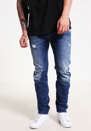 Vaqueros de mezclilla en un azul desgastado con acentos desgastados, de corte ajustado, desgastados en las rodillas, combinados con zapatillas blancas y una camiseta negra.