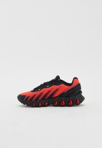 Baskets de sport avec une tige en mesh noir et rouge, design en vagues, semelle noire et accents circulaires rouges sur la semelle. Dotées d'une languette rembourrée.