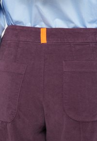 Lila hoch taillierte Hose mit glatter Textur, ausgestattet mit zwei Gesäßtaschen und einer orangen Schlaufe am Bund.