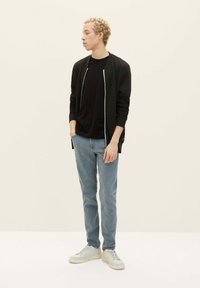 Veste zippée noire sur une chemise noire, jeans bleu clair et baskets blanches. La veste a des poignets côtelés et des poches latérales.