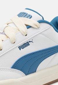 Baskets blanches avec des accents bleus, dotées d'une tige en cuir texturé, de lacets beige et d'une semelle extérieure en caoutchouc. Logo affiché sur le côté.