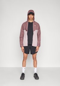 Tuulejakk heledas mauves ja hallis, kapuutsiga, millel on paelad, kombineeritud musta t-särgi ja lühikeste pükstega, kantud mustade tossude ja valgete sokkidega.