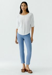 Weißes gestricktes Oberteil mit lockerer Passform und 3/4-Ärmeln, kombiniert mit enganliegenden hellblauen Hosen und braunen Plateausandalen. Einfaches und lässiges Outfit.