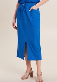 Jupe midi bleue côtelée avec poches, taille à cordon de serrage et fente à l'avant. Le tissu est texturé et léger, associé à des sandales à talons avec lanières.