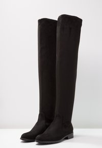 Botas negras de ante por encima de la rodilla, con una textura suave, puntera redondeada y tacones planos, presentando un diseño sin costuras y sin hardware visible.