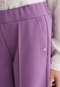 Tuvplāns uz personu, kas valkā violeto augsta jostasvietas bikses ar mazu saules logo tagu un baltu blūzi ar violetu izšuvumu aprocē.