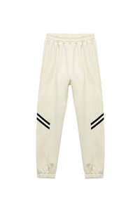 Cremefarvede sweatpants med mesh-paneler i siden, sorte striber på siderne og elastisk talje. Blød tekstur og tapered pasform.