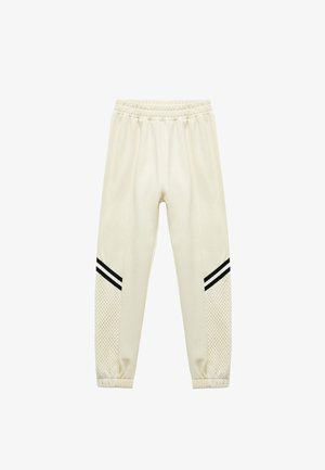 Cremefarbene Jogginghose mit seitlichen Netzpaneelen, schwarzen Streifen an den Seiten und einem elastischen Bund. Weiche Textur und schmal zulaufender Schnitt.