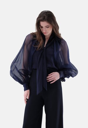 Marineblaue durchsichtige Bluse mit gepufften Ärmeln, Krawattenhals und gerafften Bündchen, über einem einfarbigen dunklen Oberteil getragen. Glatte, leichte Textur.