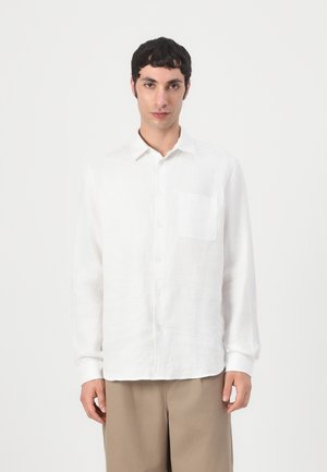 Junger Mann mit kurzen schwarzen Haaren, der ein weißes Hemd mit Knöpfen und beige Hosen trägt, steht vor einem schlichten hellen Hintergrund.