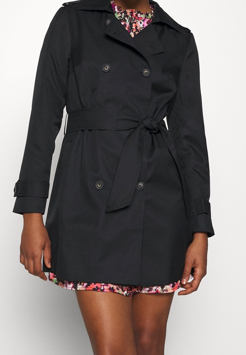 Trench-coat noir croisé avec ceinture à la taille porté sur une robe fleurie colorée, visible de la mi-cuisse aux épaules.