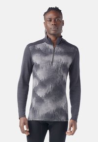 Man in een lange mouwen, kwart-rits pullover met een grijswaarden bospatroon aan de voorkant en effen donkergrijze mouwen.