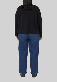 Vrouw met halflang donker haar, gekleed in een zwart shirt met lange mouwen, blauwe spijkerbroek en zwarte schoenen, staand met de rug naar de kijker op een grijze achtergrond.