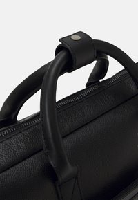 Sac à main en cuir noir avec un extérieur texturé, deux poignées robustes avec un accent bouton et une fermeture éclair en haut.