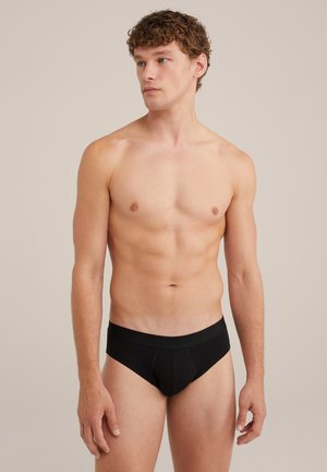 Culotte in cotone nero con una fascia larga in vita, caratterizzate da una texture liscia e una tasca frontale sagomata. Il modello è in piedi in un ambiente neutro.