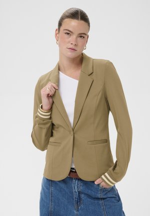 Mladá žena so zviazanými vlasmi v zadnej časti, oblečená v hnedom saku s pruhovanými manžetami, bielom topu a modrej džínovej sukni, pózuje pred jednoduchým pozadím.