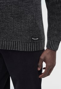 Gros plan d'une main à la peau foncée à côté d'un pull en maille gris foncé et noir avec le logo Volcom près de l'ourlet, associé à un pantalon noir.