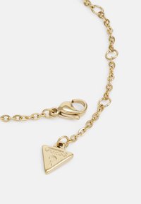 Bracelet en chaîne dorée avec un charme triangulaire portant le texte embossé "GUESS". Fermoir en forme de perroquet avec une finition brillante et des maillons espacés uniformément.