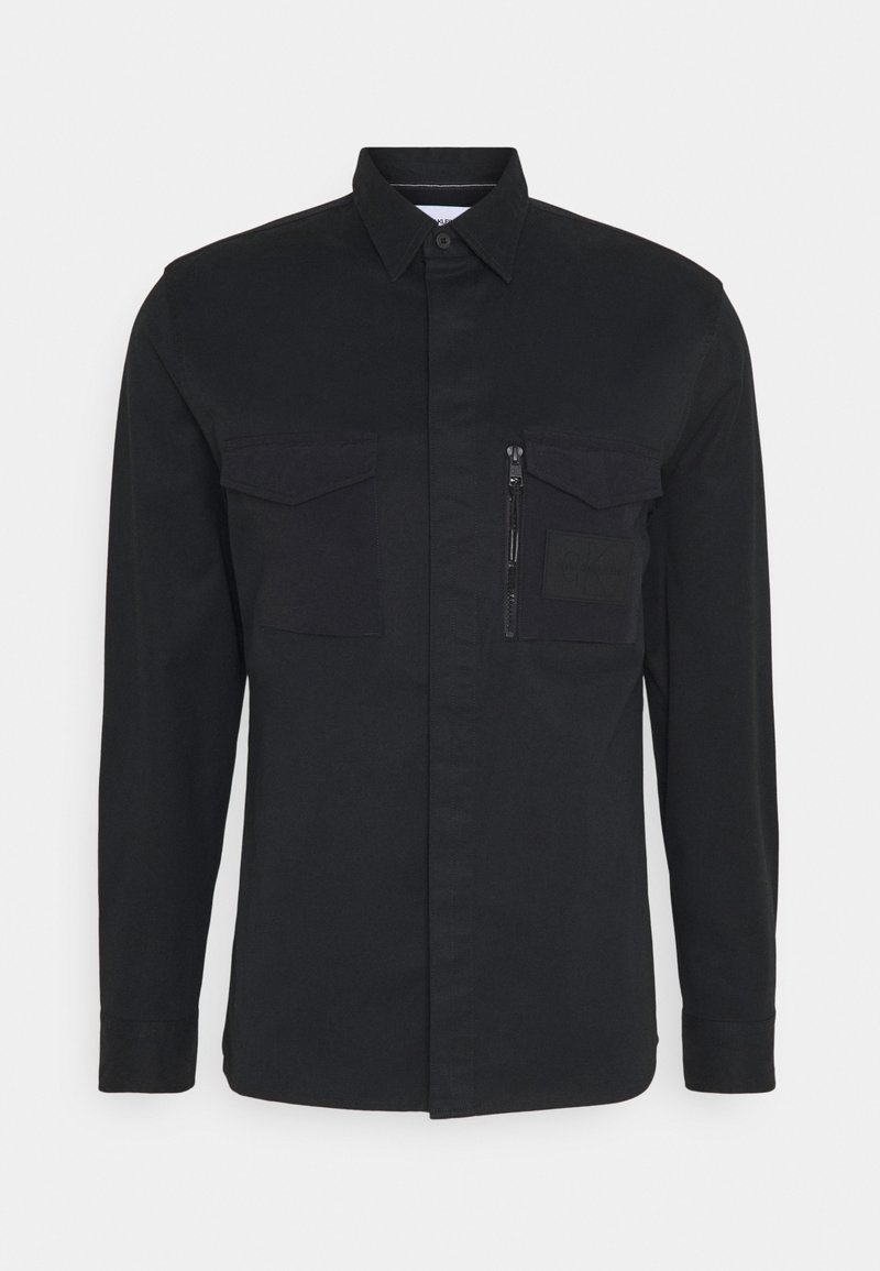 Camicia nera con bottoni, dotata di due tasche sul petto, una tasca con zip e un dettaglio a toppa. Realizzata in un tessuto resistente con una superficie liscia.