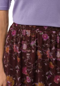 Gros plan sur une personne portant un haut lilas rentré dans une jupe florale sombre ornée de motifs floraux roses, orange et jaunes.