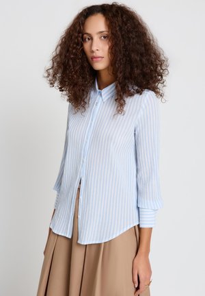 Femme aux cheveux bouclés portant une chemise à rayures bleu clair et blanc boutonnée et une jupe plissée beige, debout devant un fond uni.