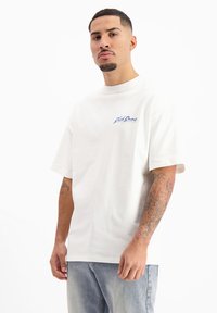 Witte katoenen t-shirt met korte mouwen, voorzien van een blauw logo op de linkerborst. De stof lijkt zacht en heeft een relaxte pasvorm.