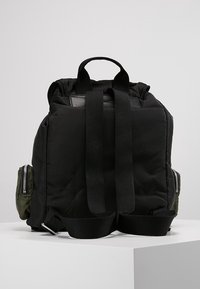Sac à dos rembourré noir avec des bretelles réglables noires, une poignée supérieure et des poches latérales vertes avec des zippers. Texture lisse, design minimaliste.
