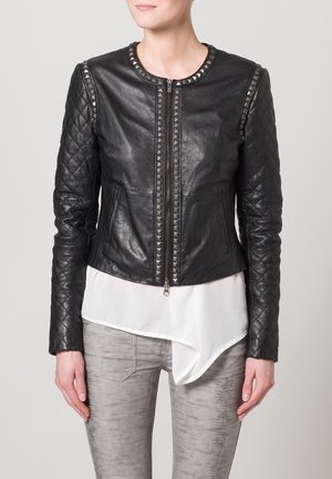 Schwarze Lederjacke mit cropped Schnitt, gesteppten Ärmeln, Nietenkragen, Reißverschluss vorne und seitlichen Taschen, getragen über einem weißen asymmetrischen Oberteil.