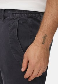 Dark navy cotton pants with an elastic waistband, side pockets, and a small logo embroidery on the left side. Gold bracelet on the wrist.