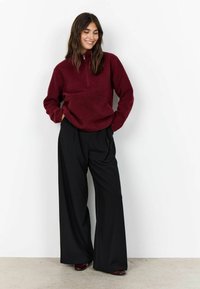 Vrouw in een bordeauxrode fleece pullover en brede zwarte broek, staand met handen in de zakken, glimlachend tegen een witte muur.