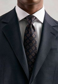 Costum în nuanță de marină, cu un finisaj texturat, cămașă albă și cravată cu model geometric în nuanțe de albastru și maro.