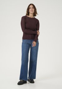 Pull à manches longues bordeaux avec un design en tricot texturé, associé à un jean bleu à jambes larges et des chaussures à plateforme noires, se tenant devant un fond uni.