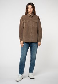 Bruine corduroy shirtjack met twee borstzakken, kraag, knoopsluiting aan de voorkant en lange mouwen; gedragen met blauwe jeans en witte sneakers.