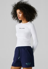 Kobieta o kręconych włosach w białej bluzce z długim rękawem Red Bull Racing oraz granatowych szortach z logo Red Bull Racing.