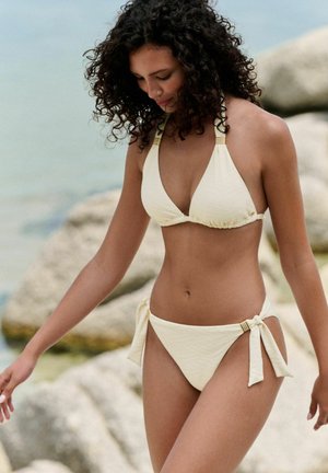 Frau mit lockigem Haar, die einen cremefarbenen Bikini mit seitlichen Bändern trägt und nahe der felsigen Küste und ruhigen See entlanggeht.